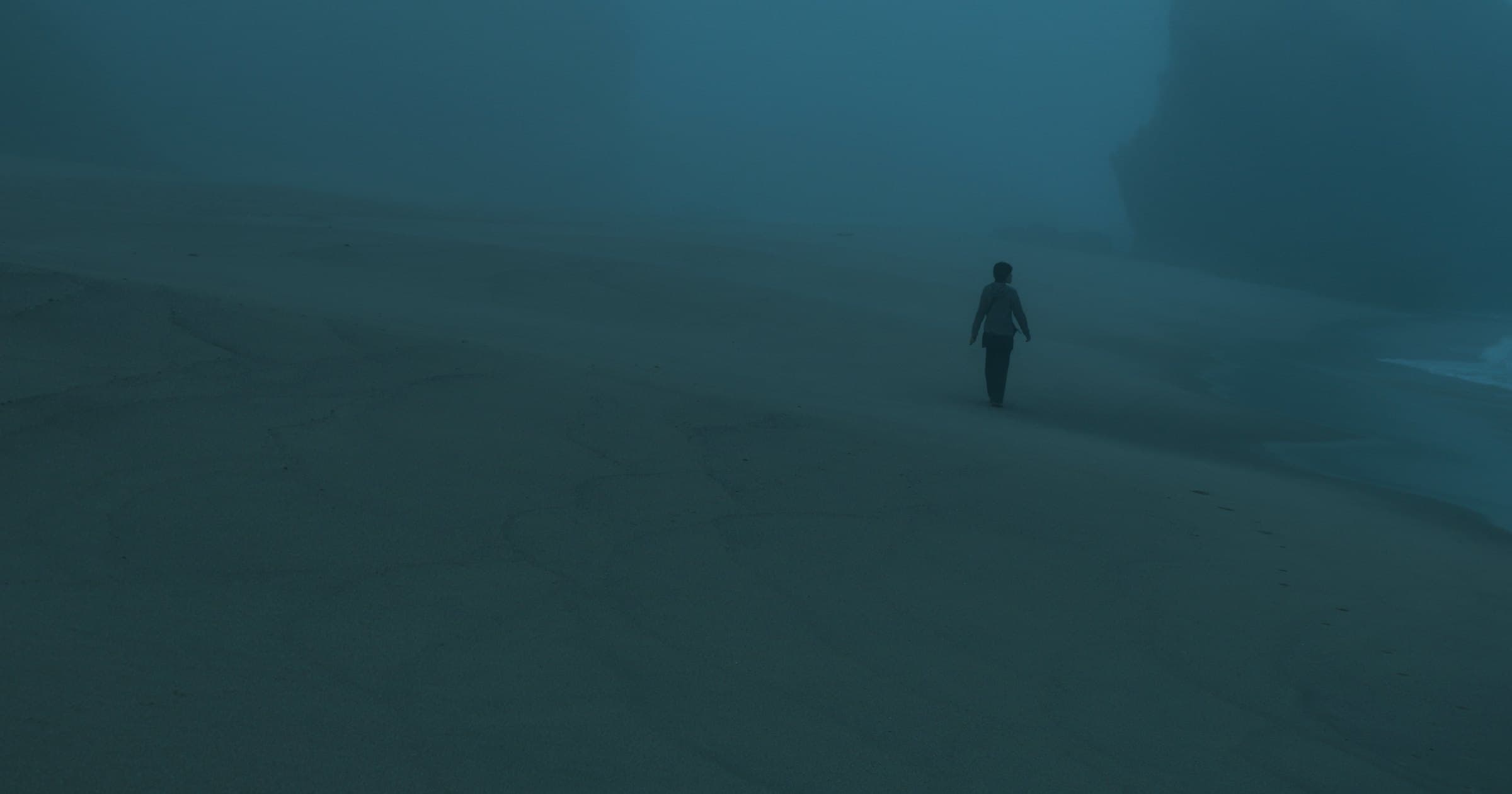 A lone figure walking on a wide beach in heavy fog.